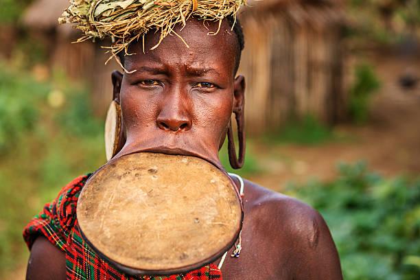 Mursi woman wearing traditional attire with a large lip plate and headdress, standing in a rural village in the Omo Valley, Southern Ethiopia